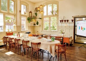 A dining area with wooden tables and chairs, large windows with plants, and a presentation screen.