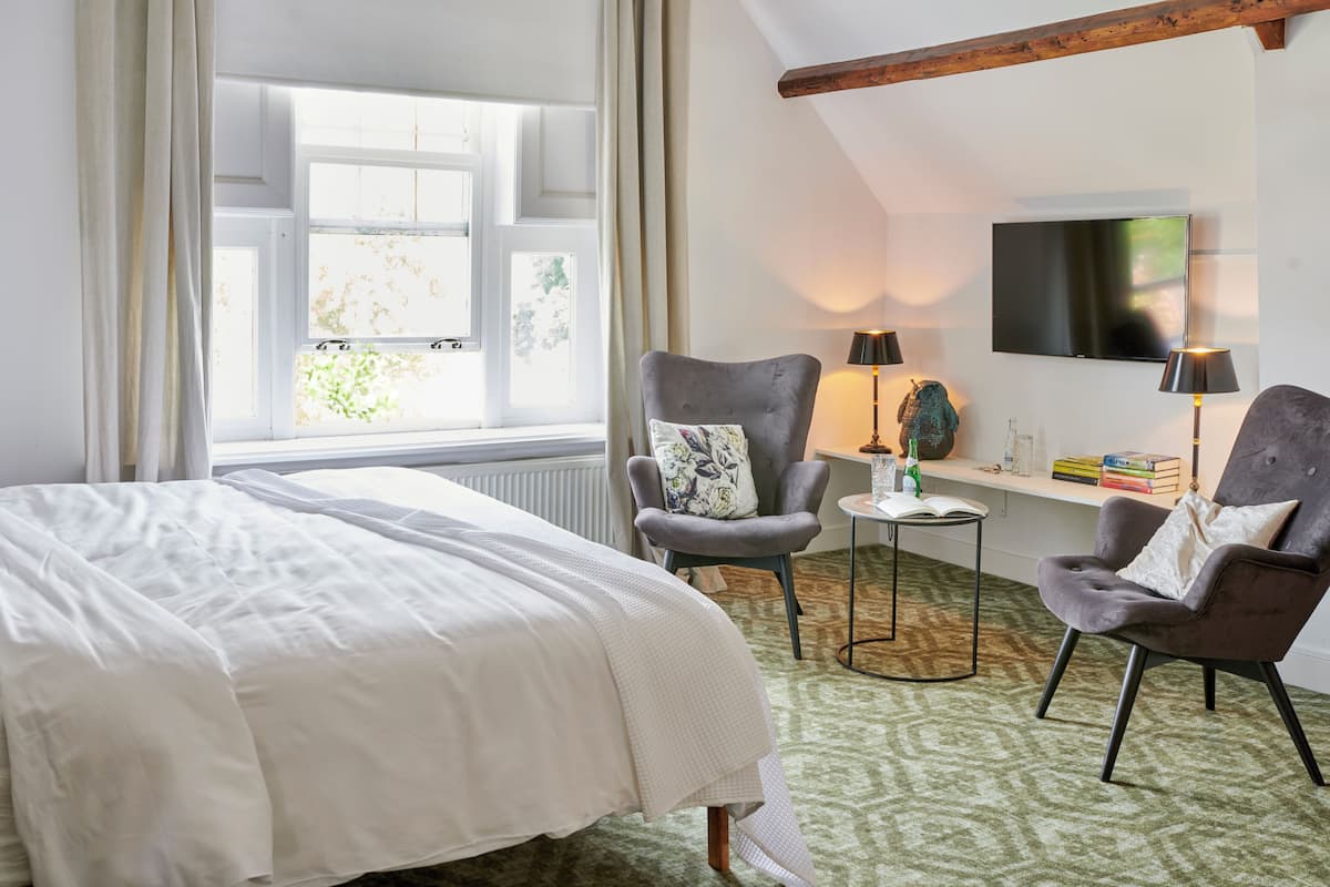 A hotel room with a large bed, two armchairs, a small table with a lamp, a television on the wall, and a window with curtains.