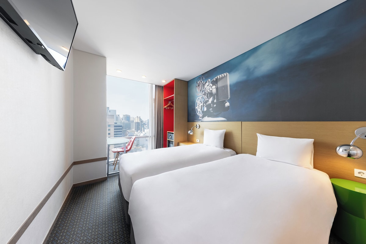 Une chambre d’hôtel avec deux lits, une vue sur la ville, un mur d’accent rouge et un grand tableau.