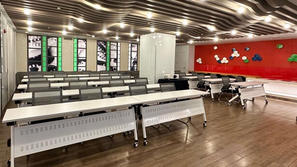 Un aula moderna con filas de mesas, una pared roja con decoraciones geométricas y un diseño de techo ondulado.