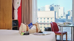 Une chambre d’hôtel avec un lit, une chaise rouge, une table avec une tasse, un appareil photo et un sac contenant des brochures de voyage.