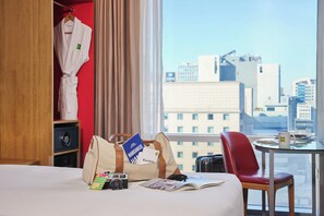 Une chambre d’hôtel avec un lit, une chaise rouge, une table avec une tasse, un appareil photo et un sac contenant des brochures de voyage.