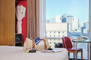 A hotel room with a bed, a red chair, a table with a mug, a camera, and a bag with travel brochures.