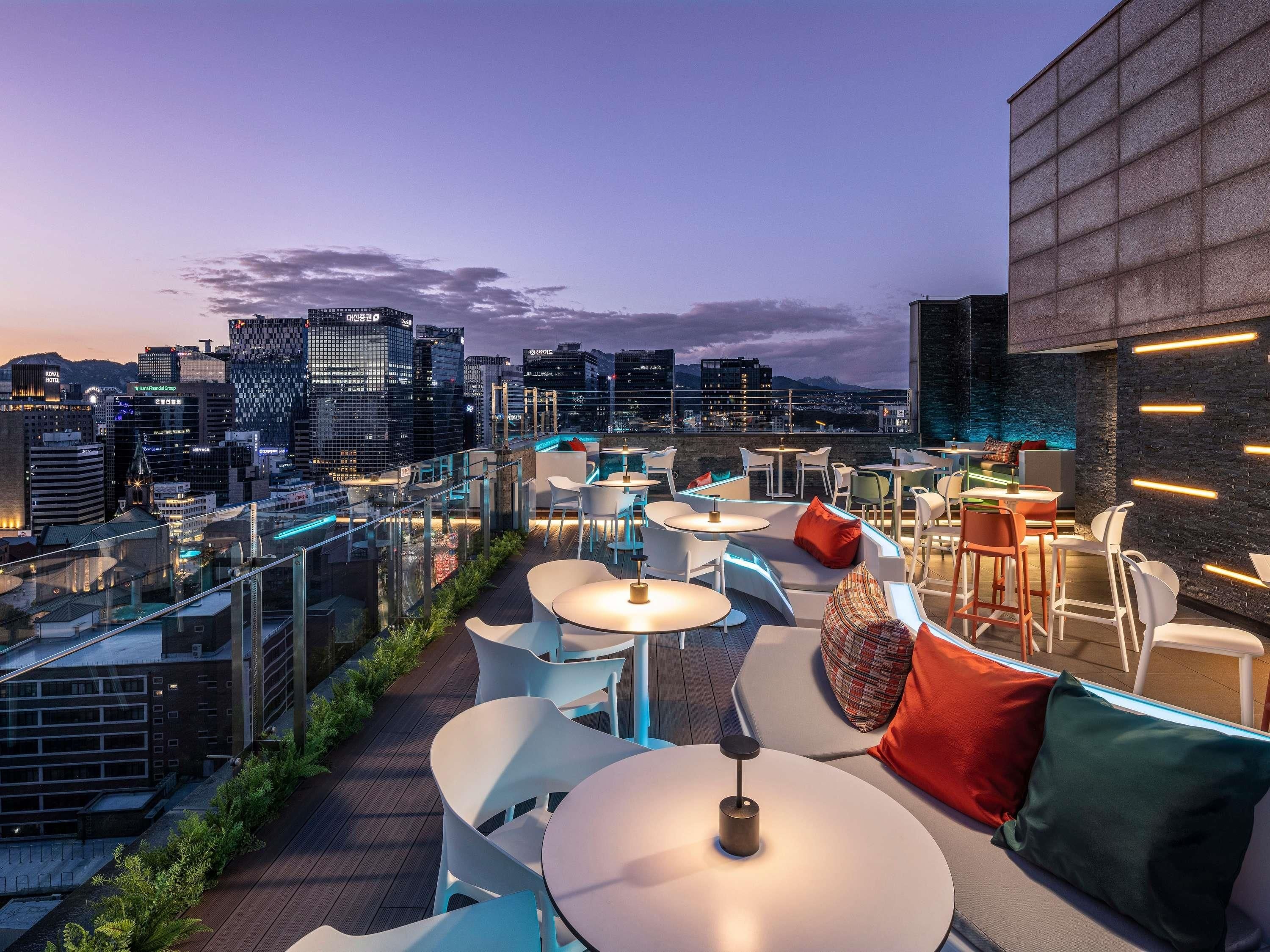 A rooftop bar with round tables and chairs, a city skyline view, and modern architecture.