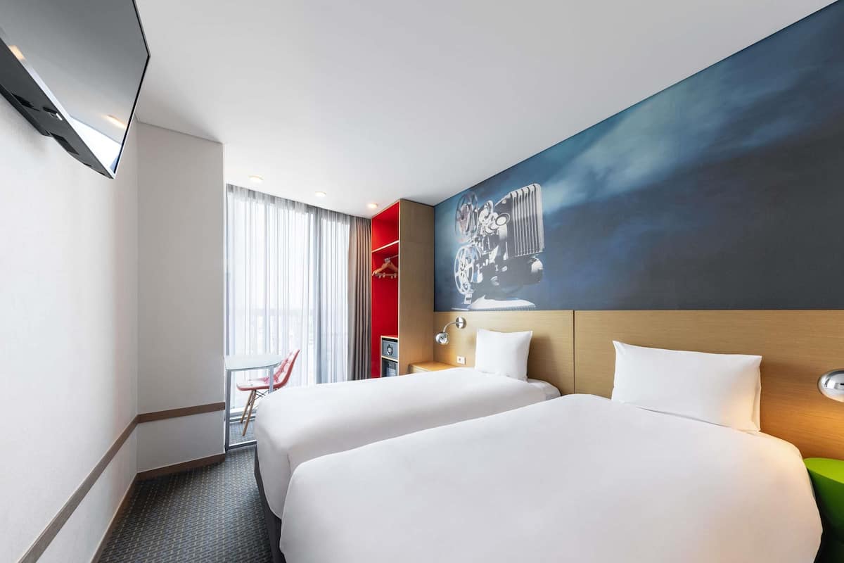 Habitación de hotel con dos camas, televisión, pared roja y ventana grande.