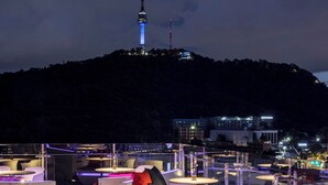 Un espacio al aire libre con mesas y sillas modernas, vista a un paisaje urbano y una torre de fondo.
