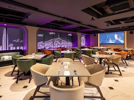 A modern restaurant with a variety of seating options, including booths and tables, and a large digital screen displaying a cityscape.