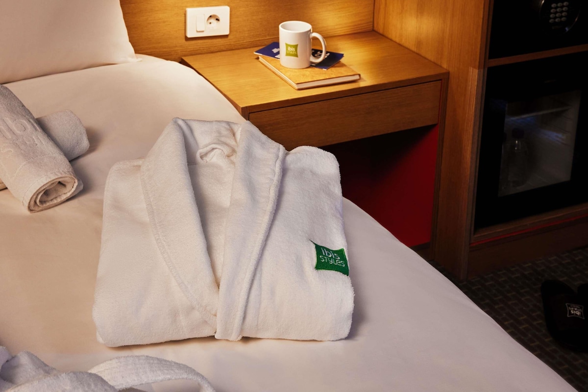 Una habitación de hotel con una cama bien hecha, una mesita de noche con un libro, una taza y un control remoto.