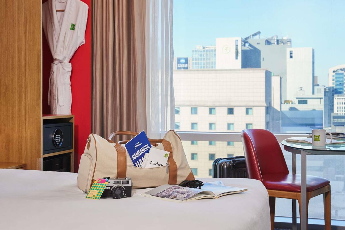 Una habitación de hotel con una cama, una silla roja, una mesa con una taza, una cámara y un bolso con documentos de viaje.