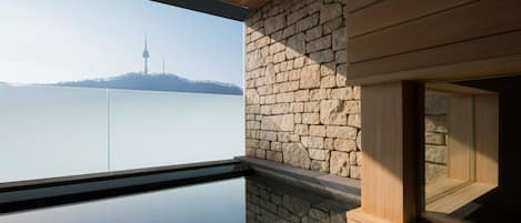 Un interior moderno con pared de piedra, techo de madera y ventana de vidrio que da a una masa de agua con una torre distante.