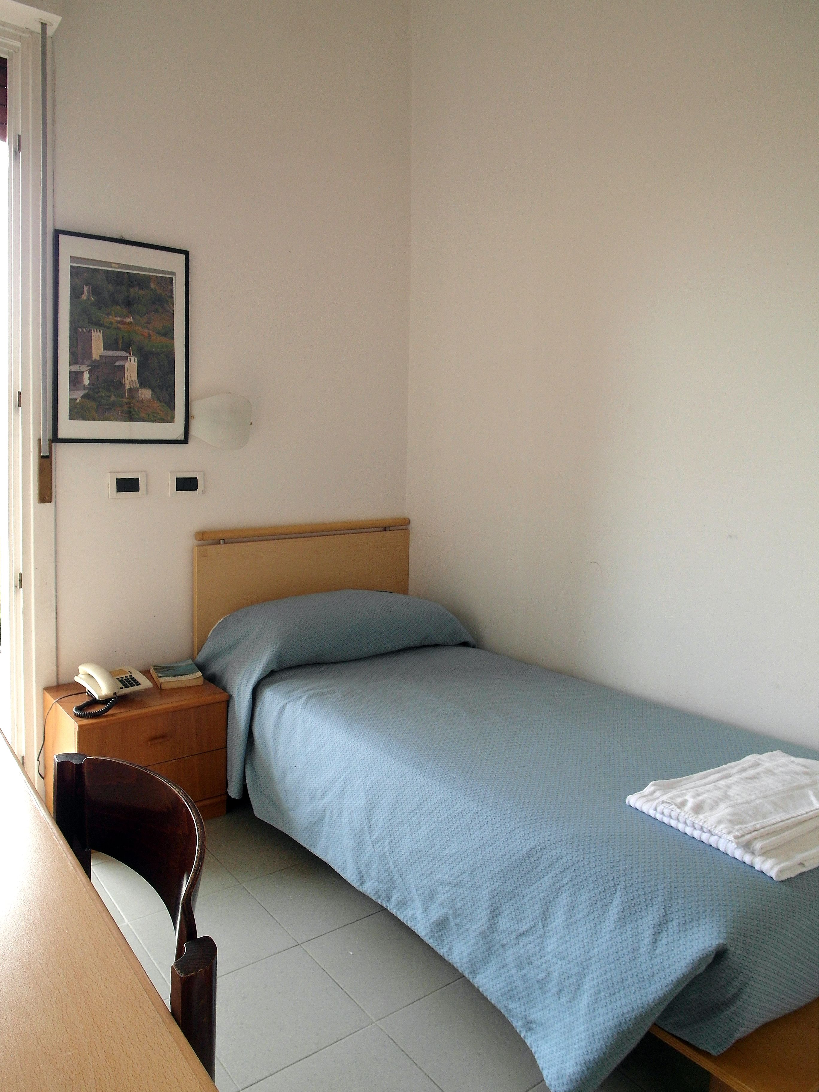 Una habitación pequeña con cama individual, cabecera de madera, mesita de noche con teléfono, un cuadro en la pared y una silla.