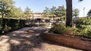 A courtyard with a gravel path, wooden planter boxes, and a view of a multi-story building with parked cars in the background. - Opera Don Calabria Casa per Ferie (Rome)