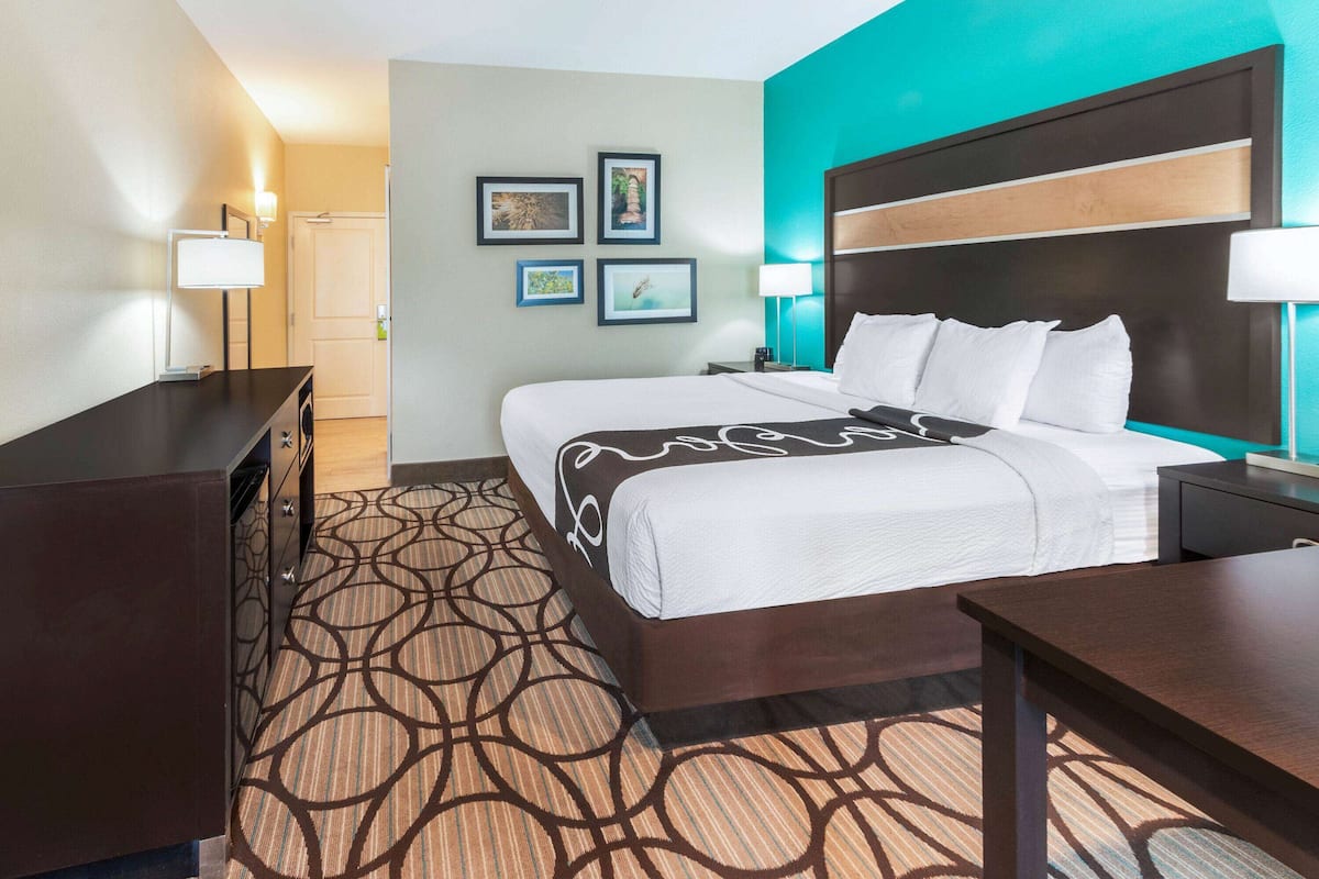 A hotel room with a large bed, a desk, a dresser, and a patterned carpet. There are wall-mounted lamps and framed pictures on the wall.