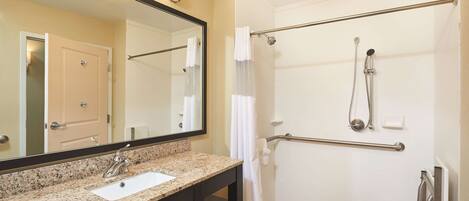 A bathroom with a granite countertop sink, a mirror, a shower with a grab bar, and a door.