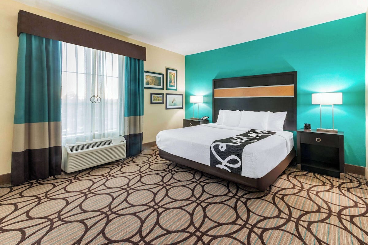 A hotel room with a bed, air conditioning unit, two bedside tables, a nightstand, and a window with curtains.
