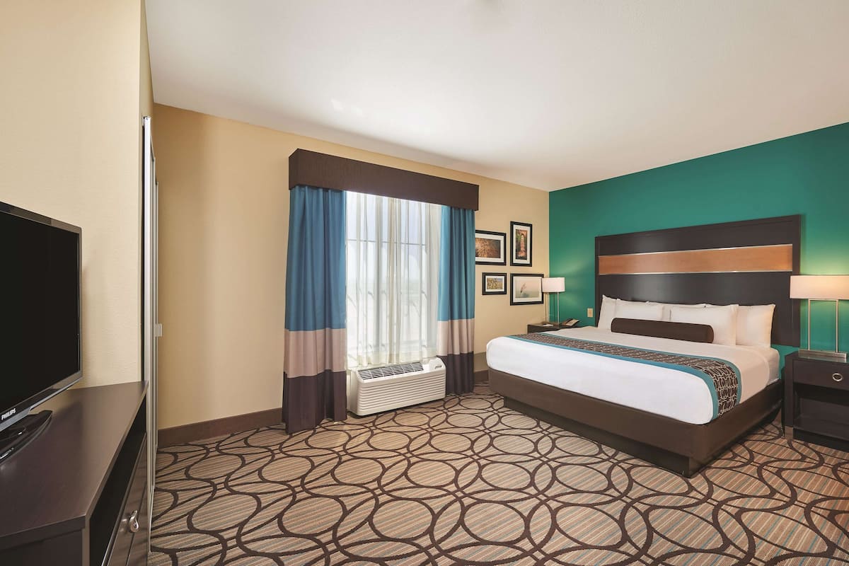 A hotel room with a bed, a television, a nightstand, a lamp, a window with curtains, and a wall-mounted air conditioner.