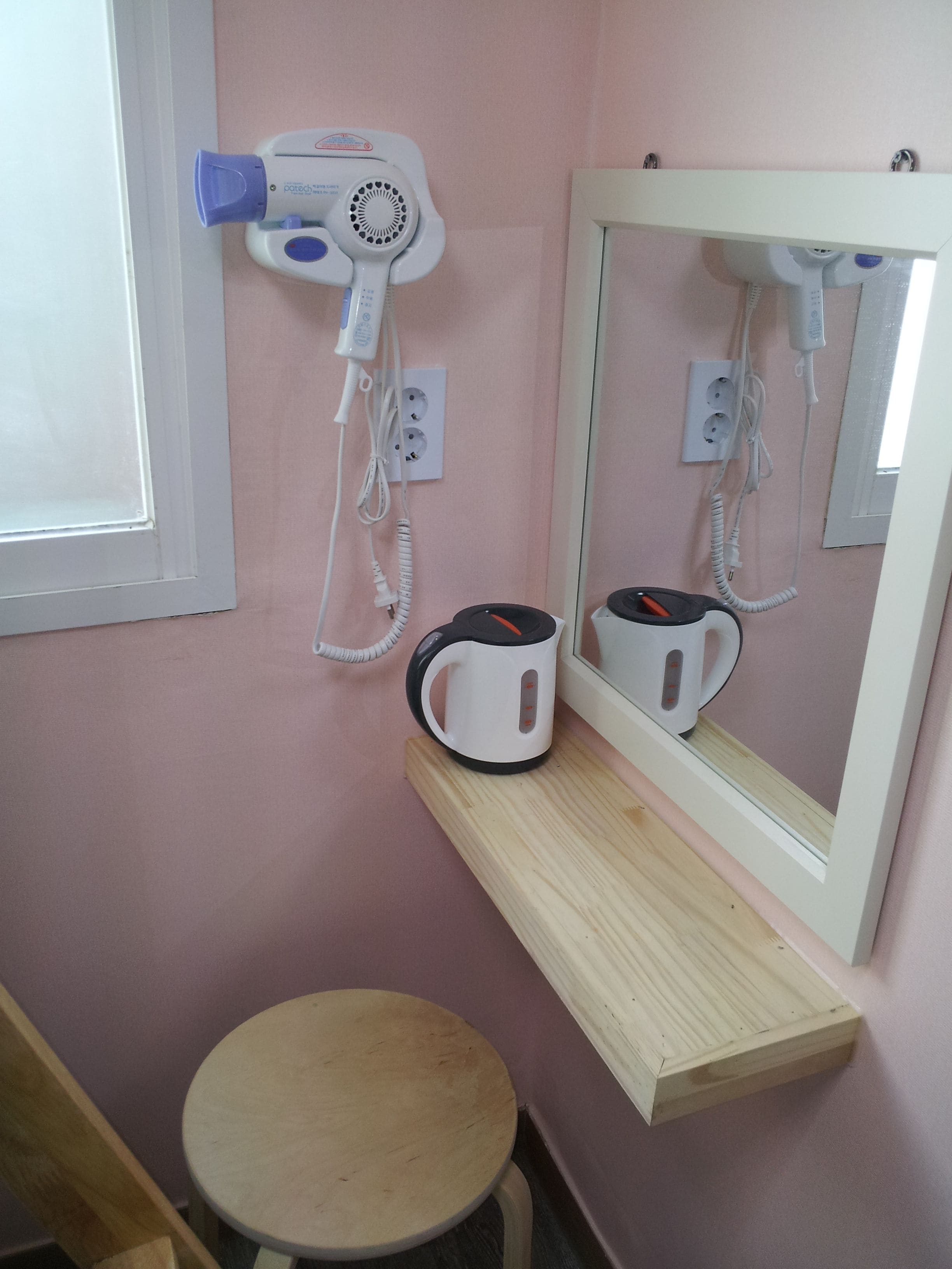 A bathroom with a pink wall, a wooden stool, a mirror, a hairdryer, and a kettle on a shelf.