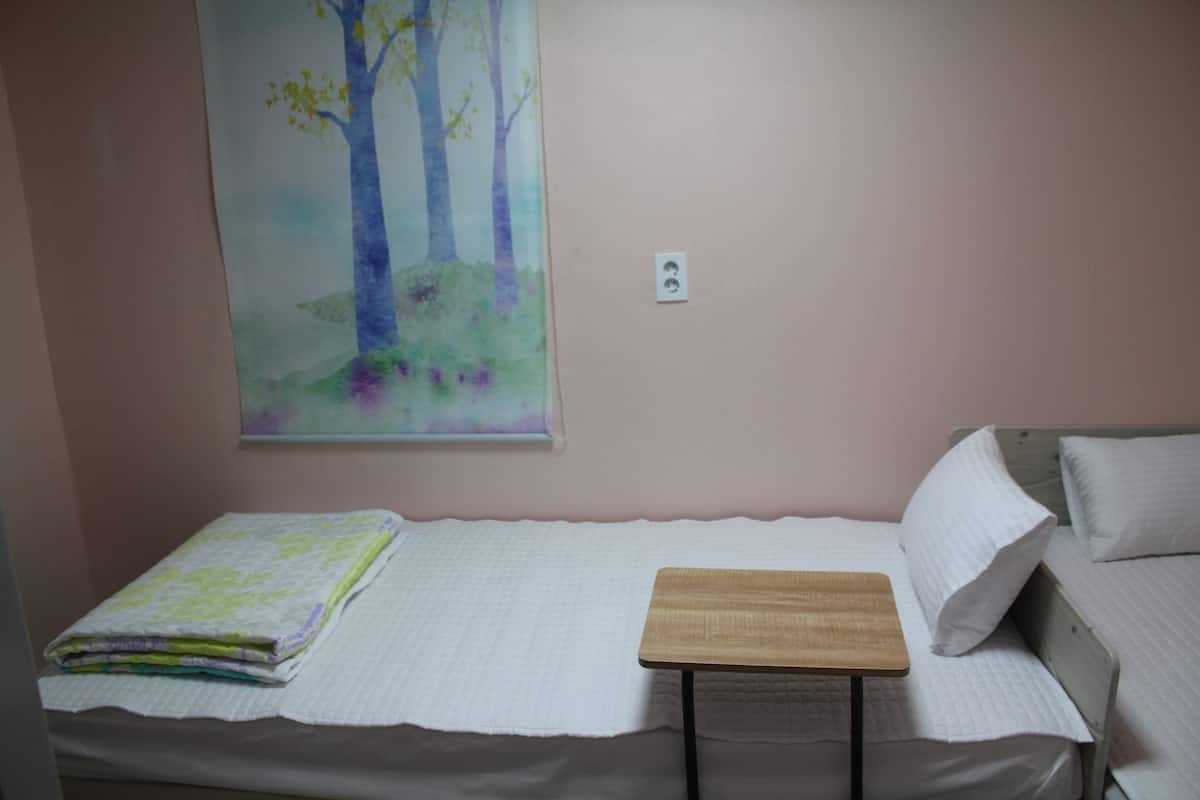 Una cama individual con colcha blanca, una mesita de madera y un cuadro de árboles colgado en la pared.