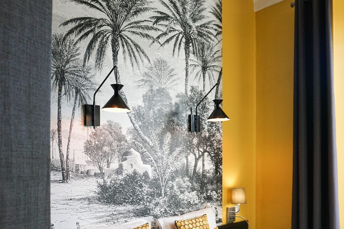 A hotel room with a large bed, two bedside tables, a wall mural of palm trees, and a dark curtain.