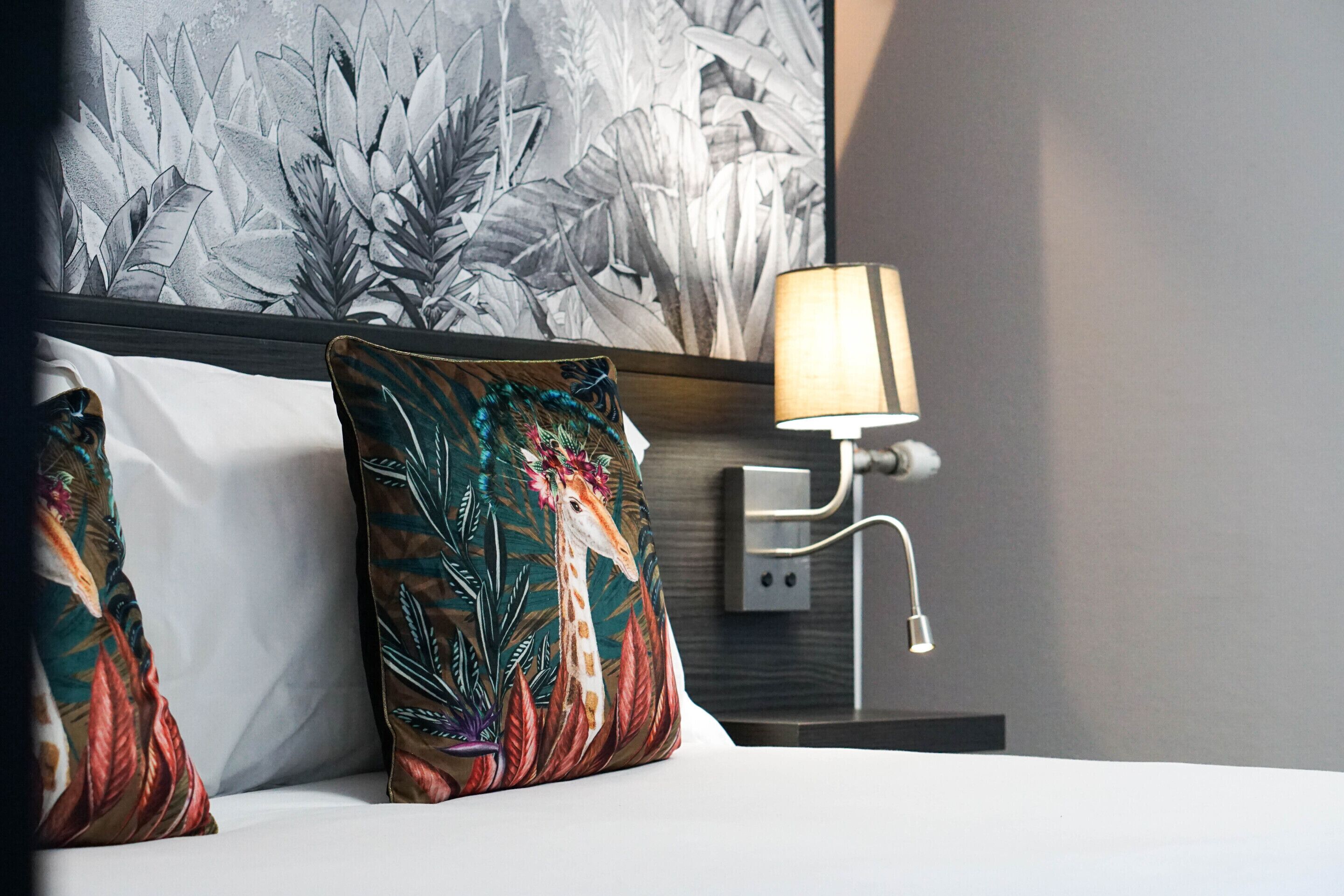Une chambre d’hôtel avec un lit, deux oreillers à motifs de girafe, une lampe fixée au mur et une grande tapisserie à motifs floraux.