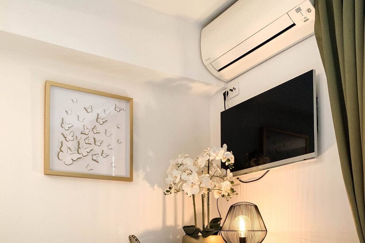 A hotel room with a bed, a desk with a chair, an air conditioner, a framed artwork, and a vase with flowers.
