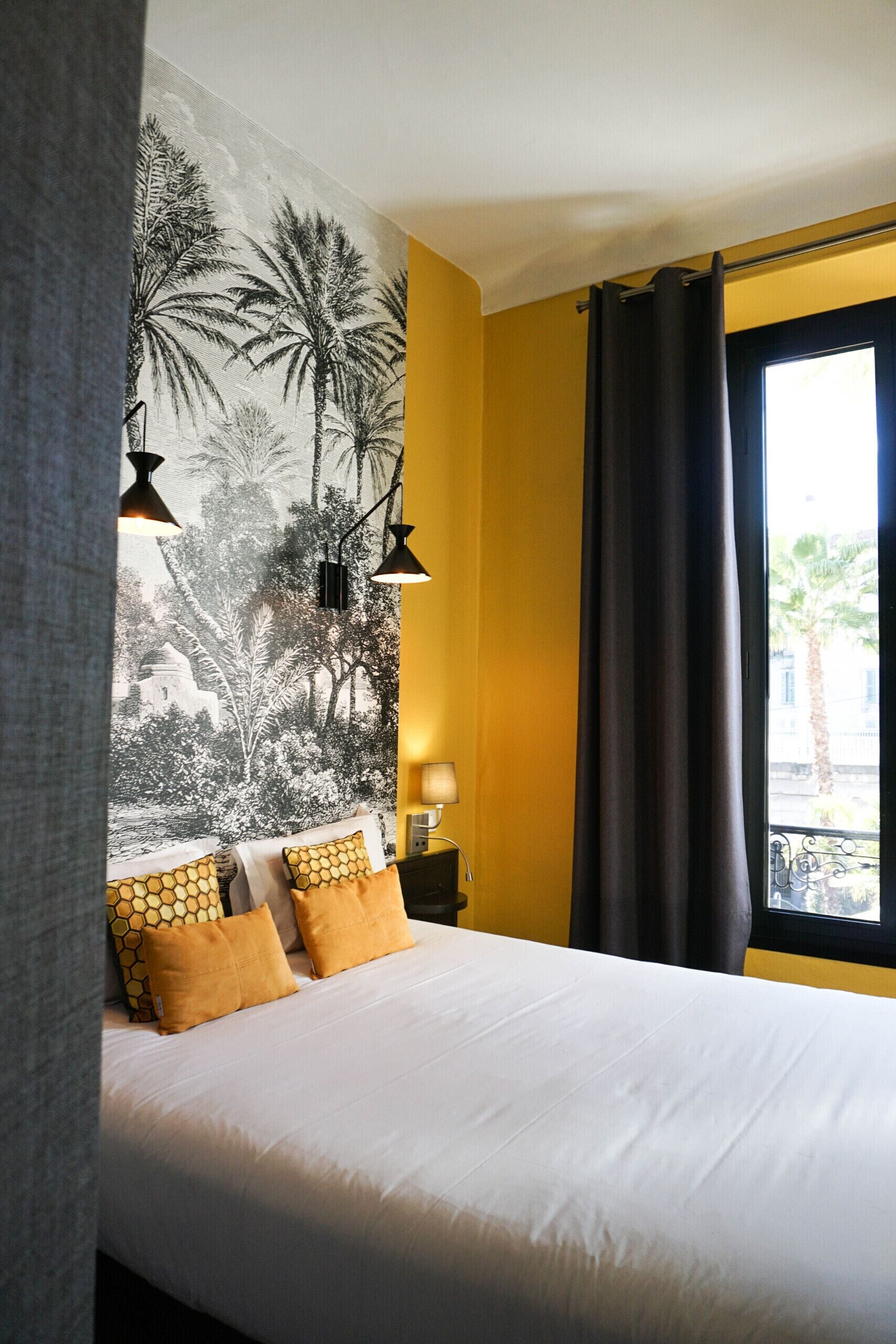 Un dormitorio con cama, un tapiz en la pared, una ventana con cortinas y una mesita de noche con lámpara.