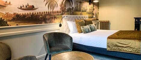 A hotel room with a large mural depicting a historical scene, a bed with a white duvet and multiple pillows, a grey armchair, a small round table, and a radiator.