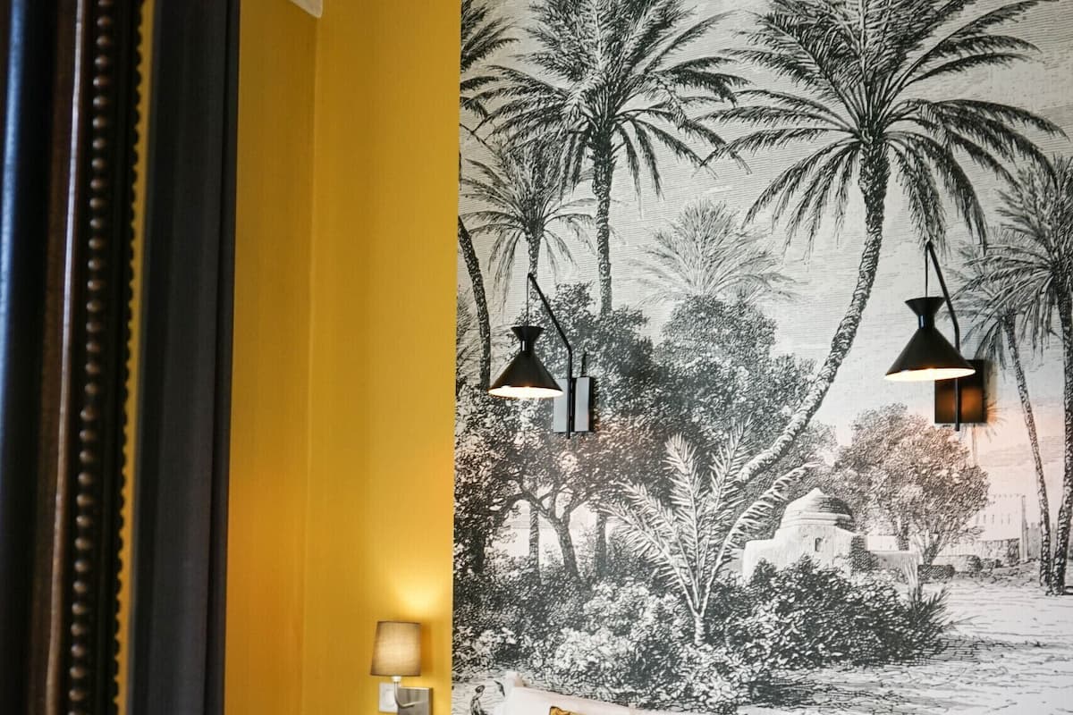 A hotel room with a bed, a nightstand, a lamp, and a wall mural of palm trees.