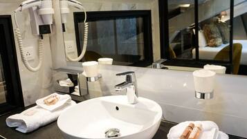 Une salle de bain moderne avec un grand lavabo blanc, un miroir et un sèche-cheveux. On y trouve des serviettes et des articles de toilette sur le comptoir.