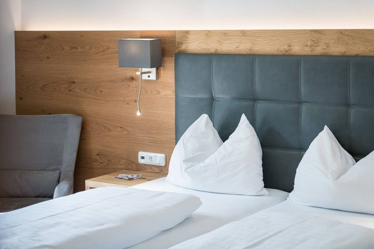 Une chambre d’hôtel avec une tête de lit en bois, un fauteuil rembourré de couleur grise, une table de chevet avec une lampe et trois oreillers blancs.