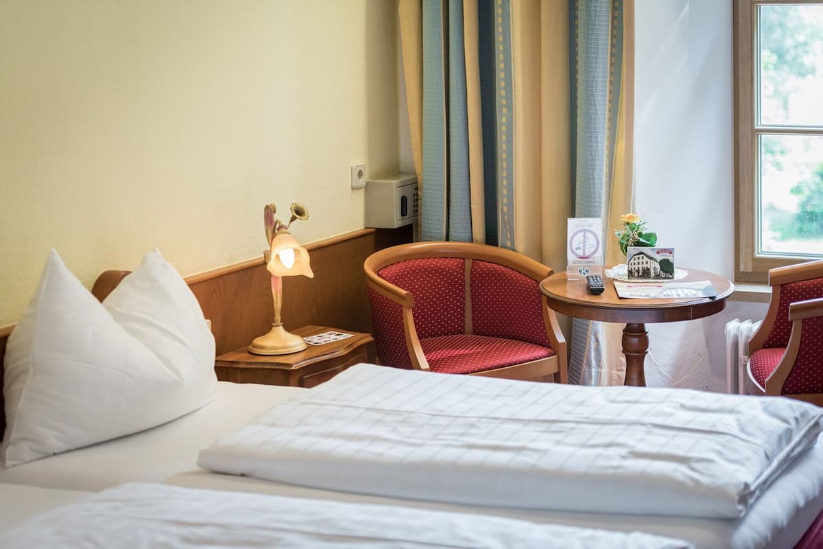 Une chambre d’hôtel avec un lit, deux chaises, une petite table avec une lampe et une fenêtre avec des rideaux.