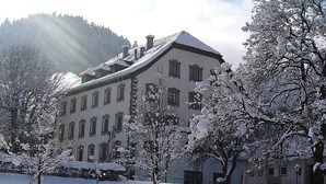 Ein großes, mehrstöckiges Gebäude mit einer symmetrischen Fassade, umgeben von schneebedeckten Bäumen und blauem Himmel.