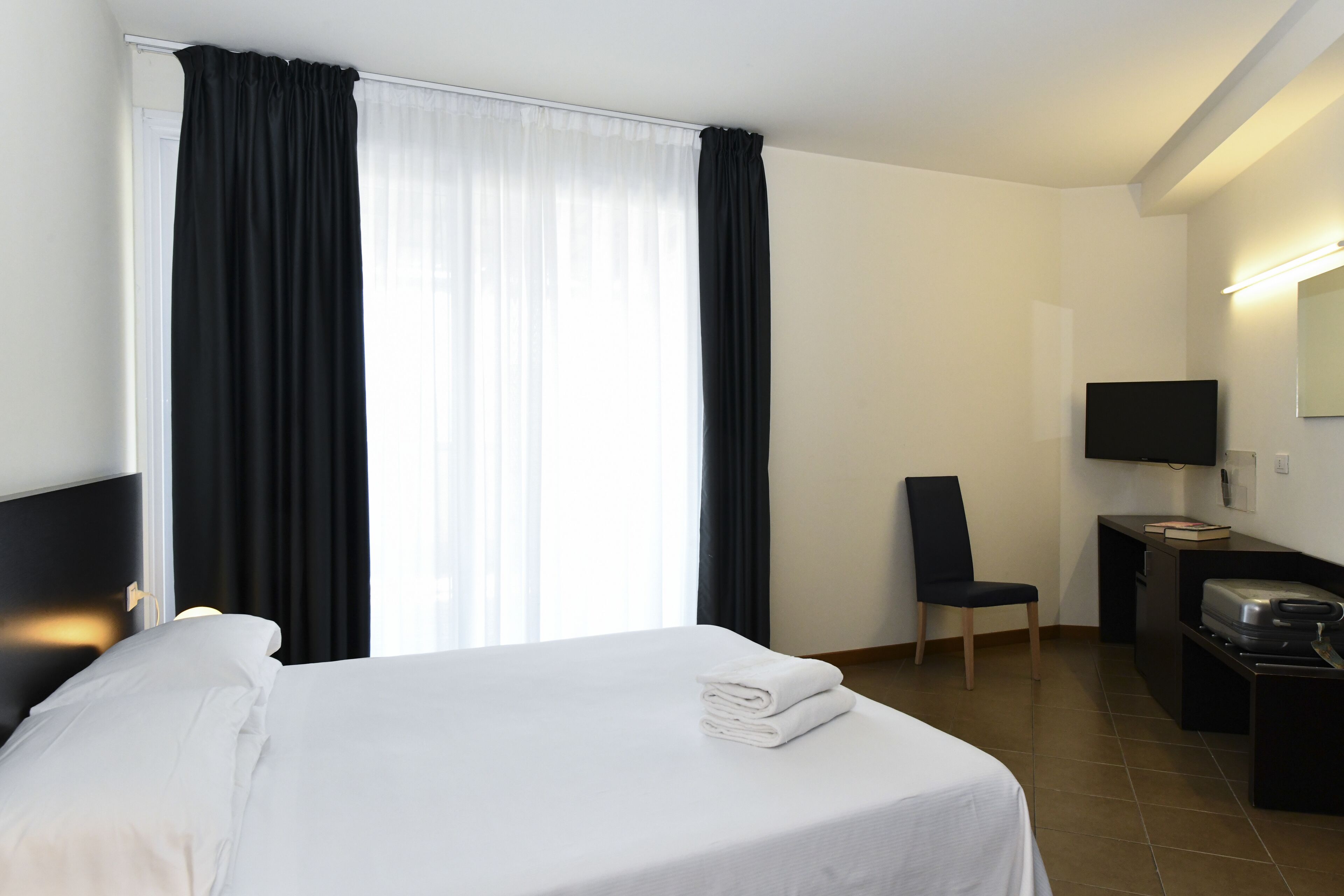 Una habitación de hotel con una cama, una silla, un escritorio con televisor y una ventana con cortinas.
