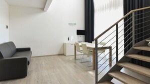 A modern living room with a sofa, a dining table with chairs, a small white cabinet, and a staircase with a railing. - Hotel Eden (Jesolo)