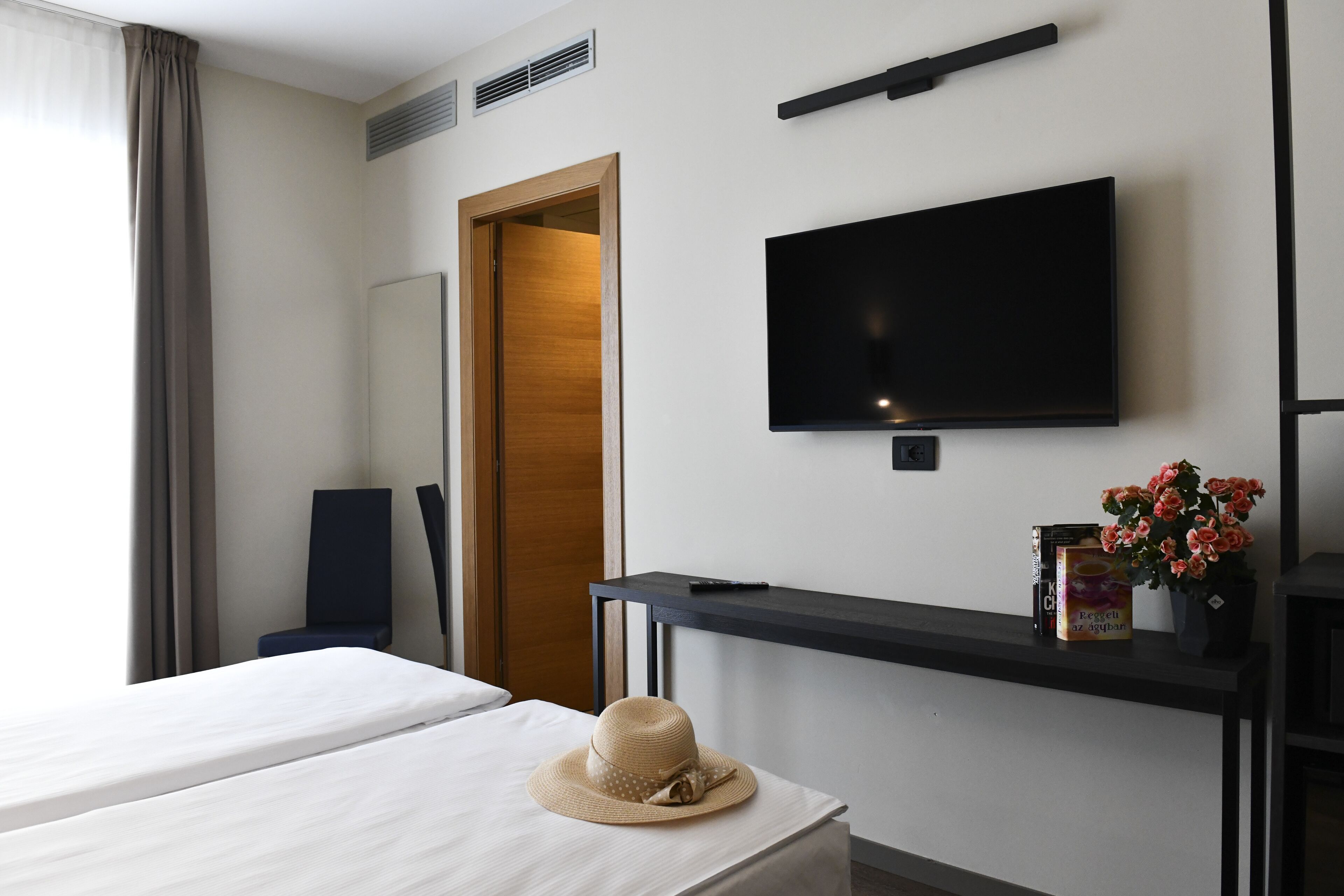 A hotel room with a bed, a television mounted on the wall, a desk with a remote control, a chair, and a potted plant.