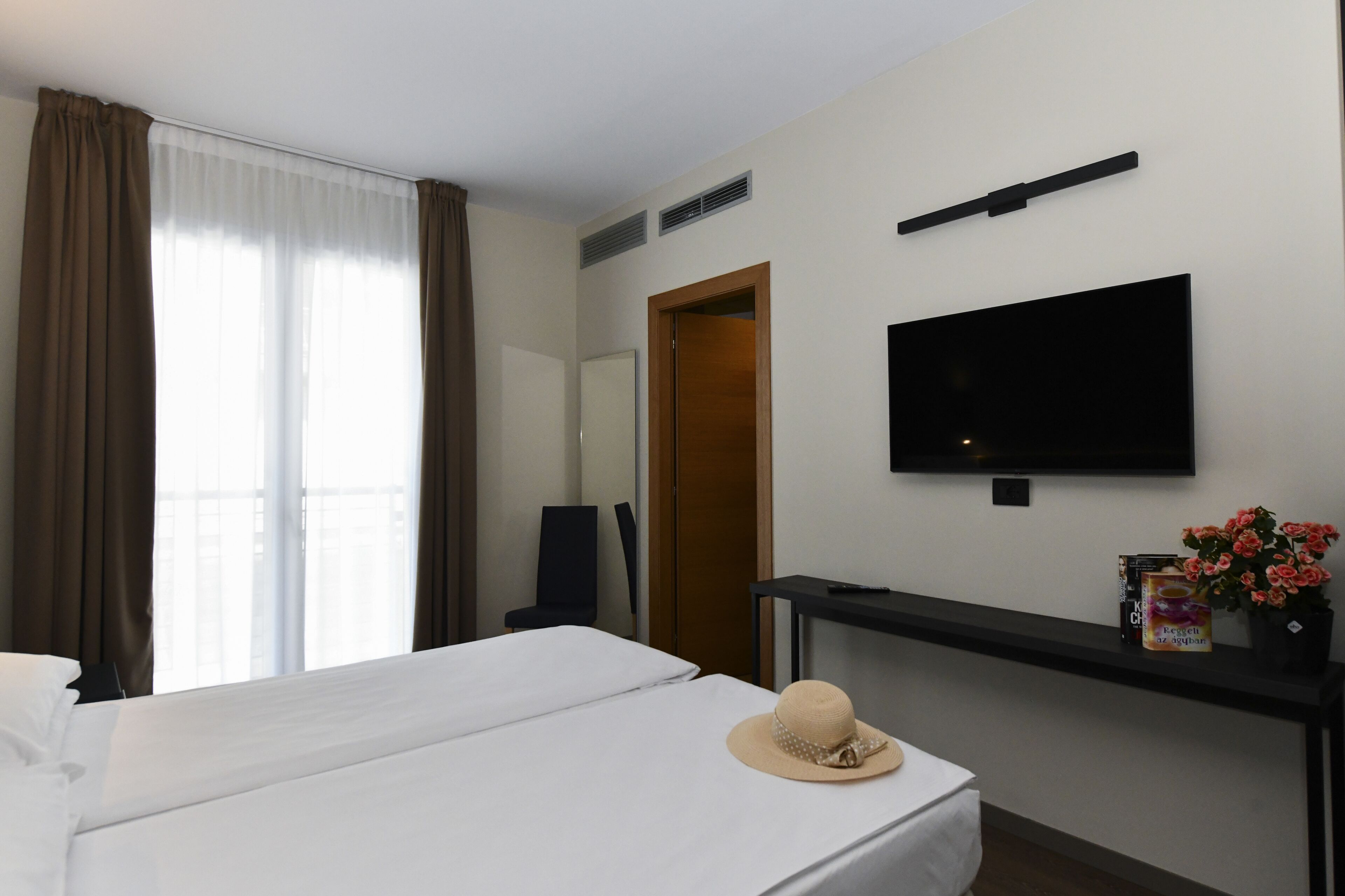 Habitación de hotel con cama, televisión montada en la pared, escritorio con computadora y ventana con cortinas.