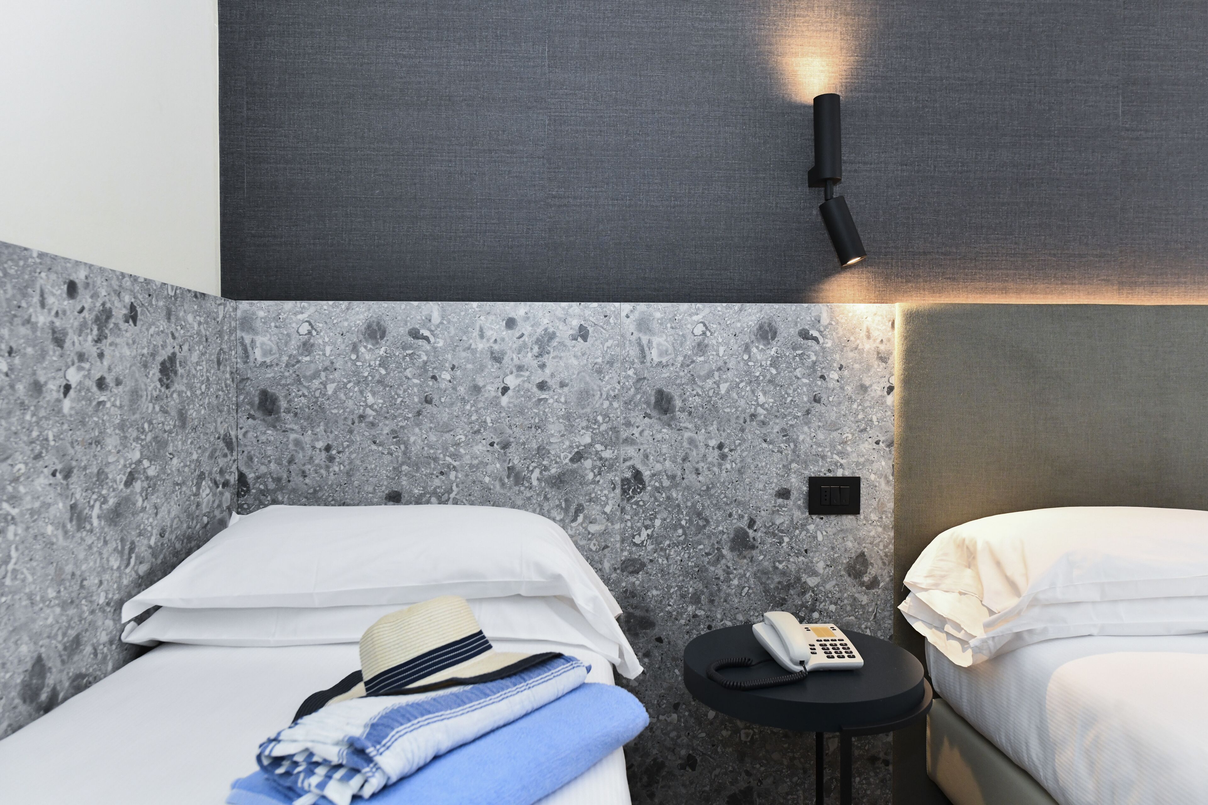 A hotel room with a textured gray wall, a bed with white bedding, a small round table with a telephone, and a wall-mounted light fixture.