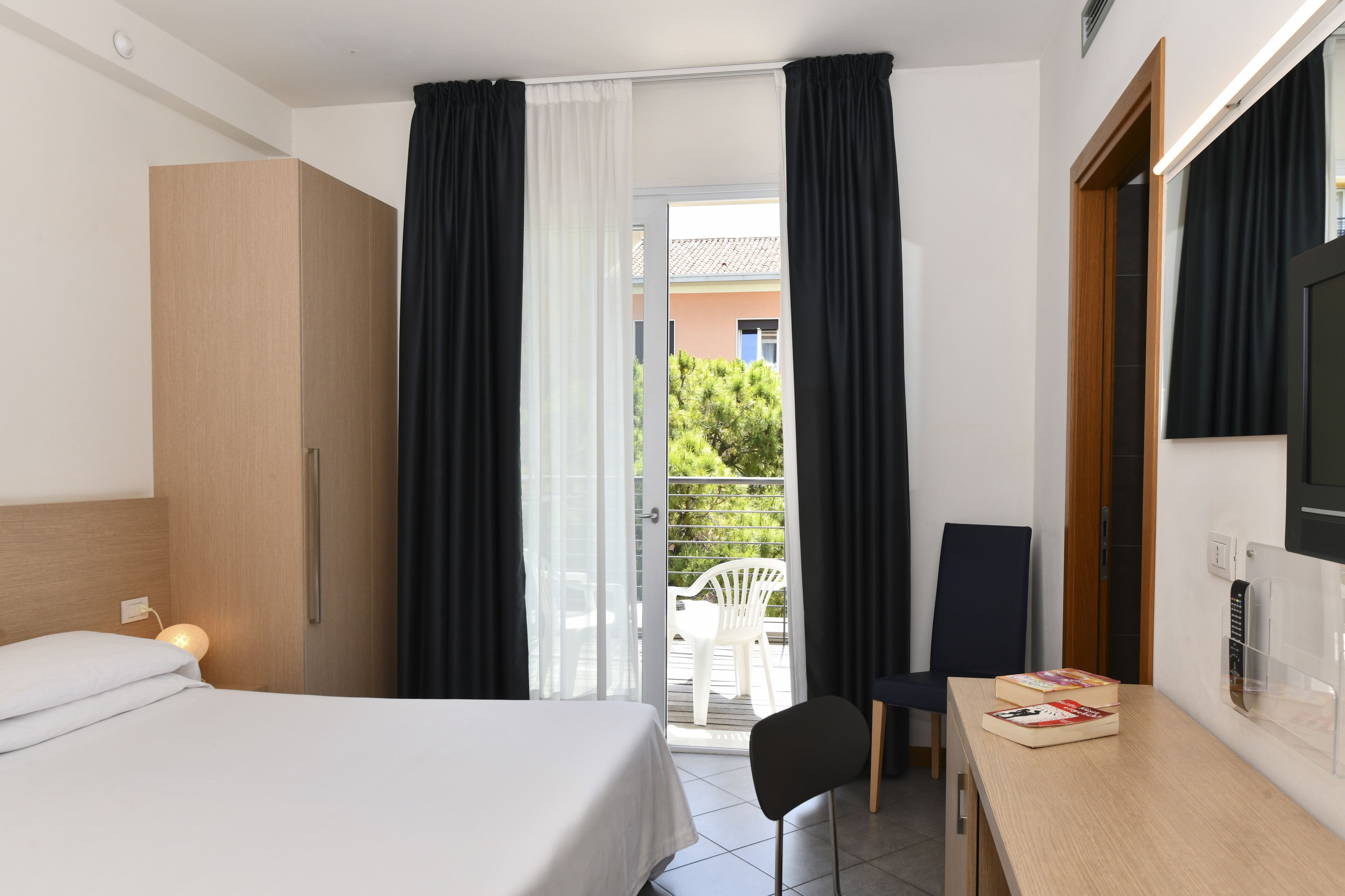 A hotel room with a bed, a desk with books, a chair, and a view of the outside through a sliding door.