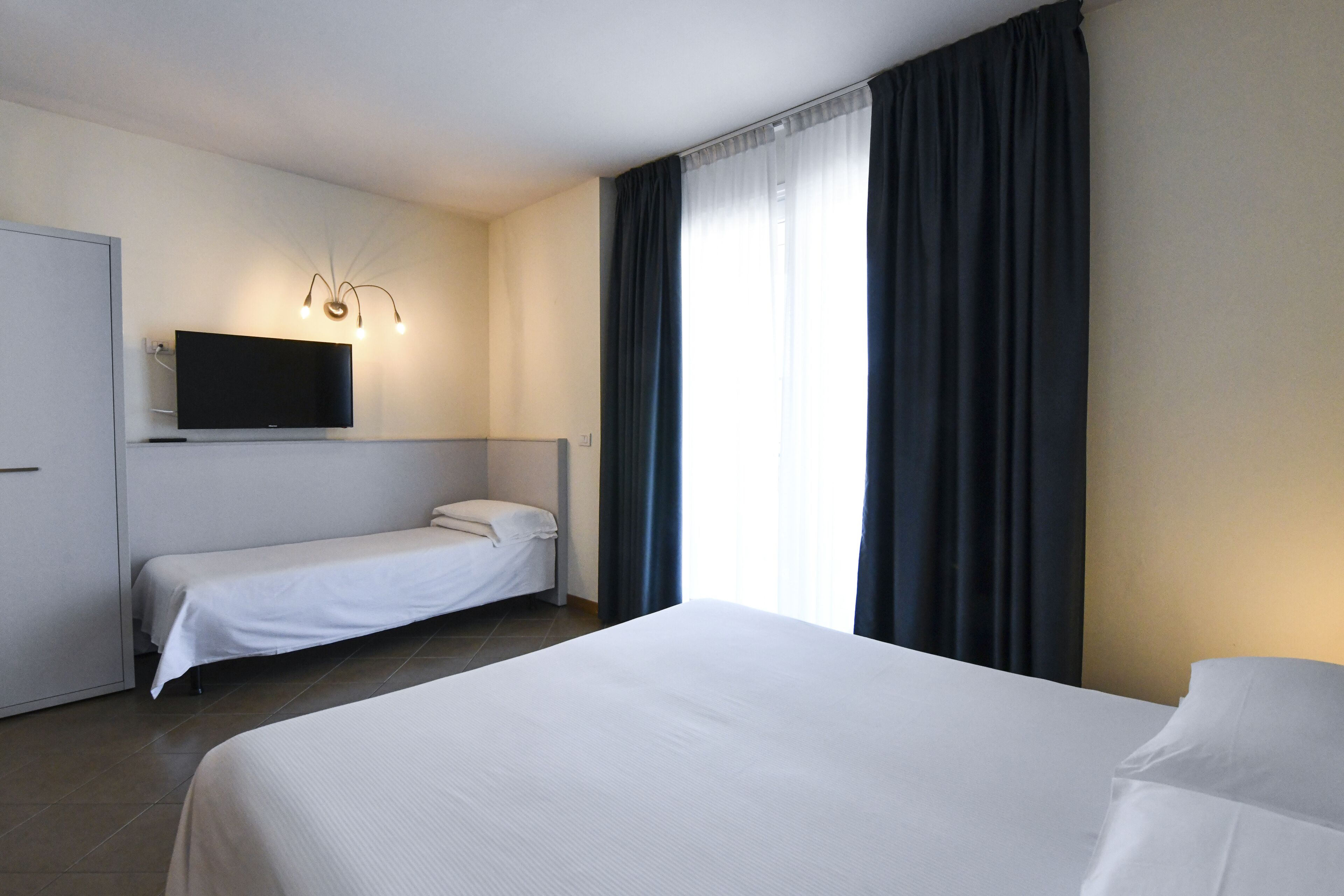 Habitación de hotel con una cama individual, una cama más pequeña, un televisor montado en la pared y una ventana con cortinas.