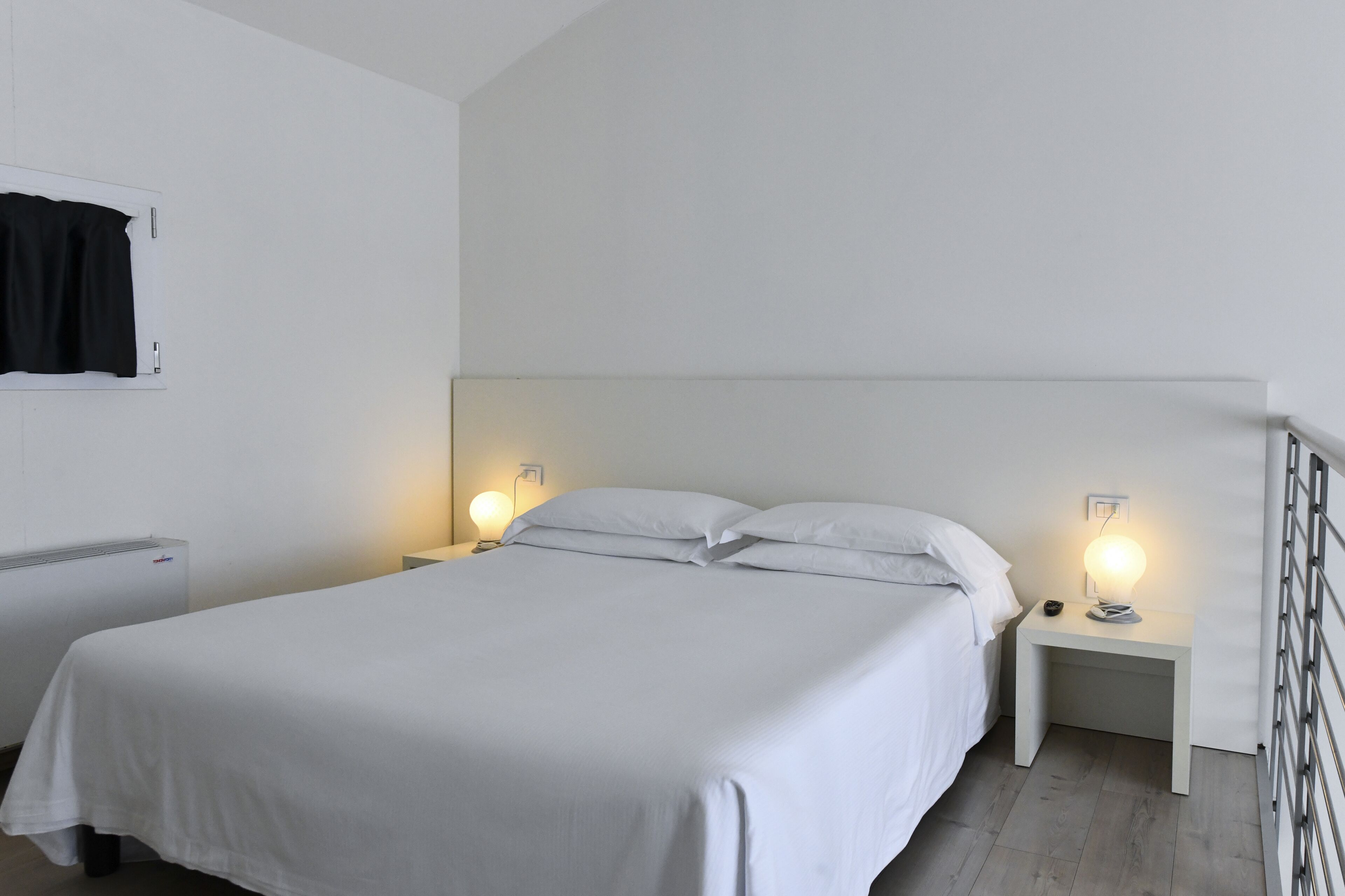Quarto individual com cama de solteiro, roupa de cama branca, duas mesas de cabeceira com luminárias, um radiador e uma cortina preta na parede.