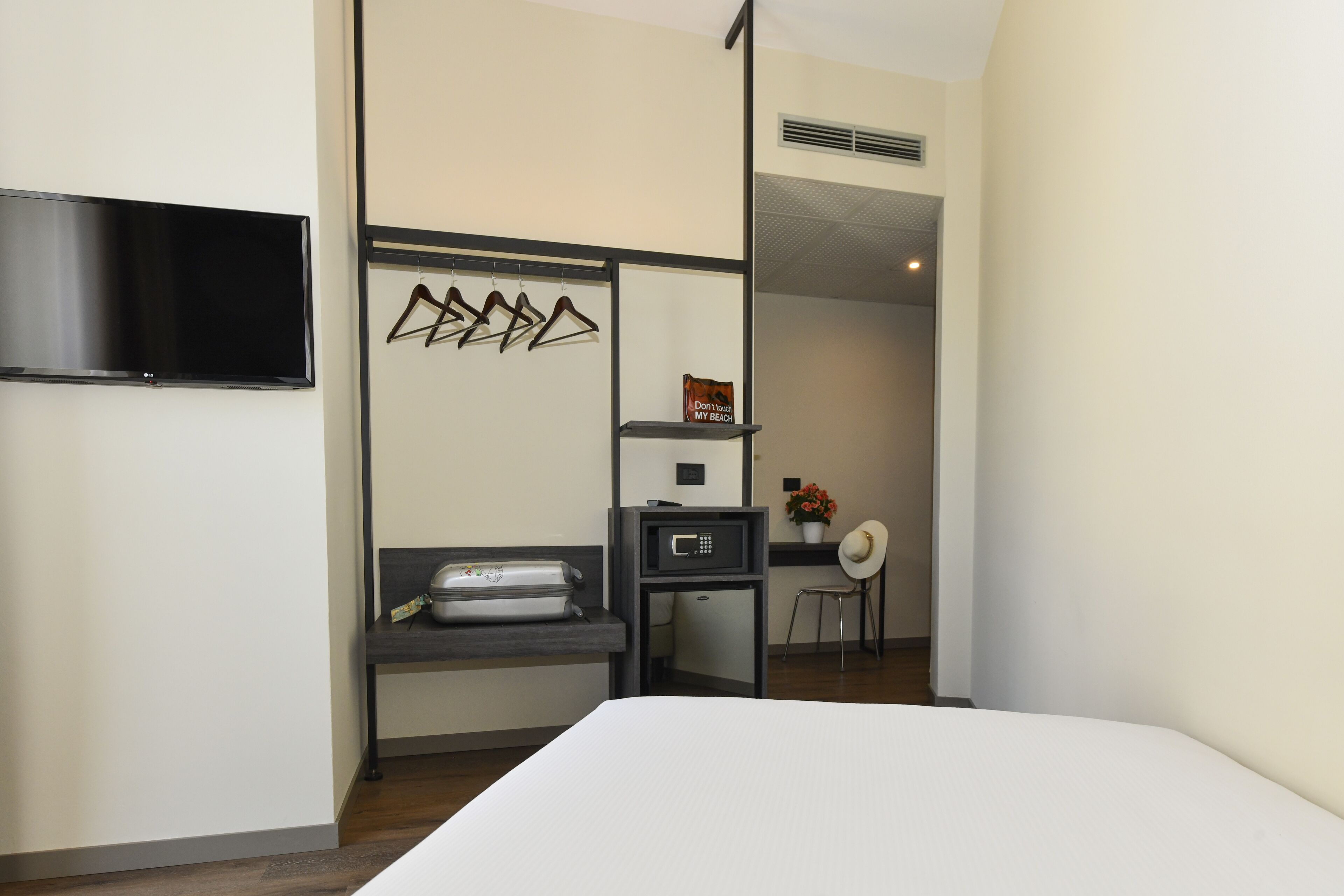 A hotel room with a bed, a flat-screen TV mounted on the wall, a small kitchenette area with a microwave, and a seating area with a chair and a small table.