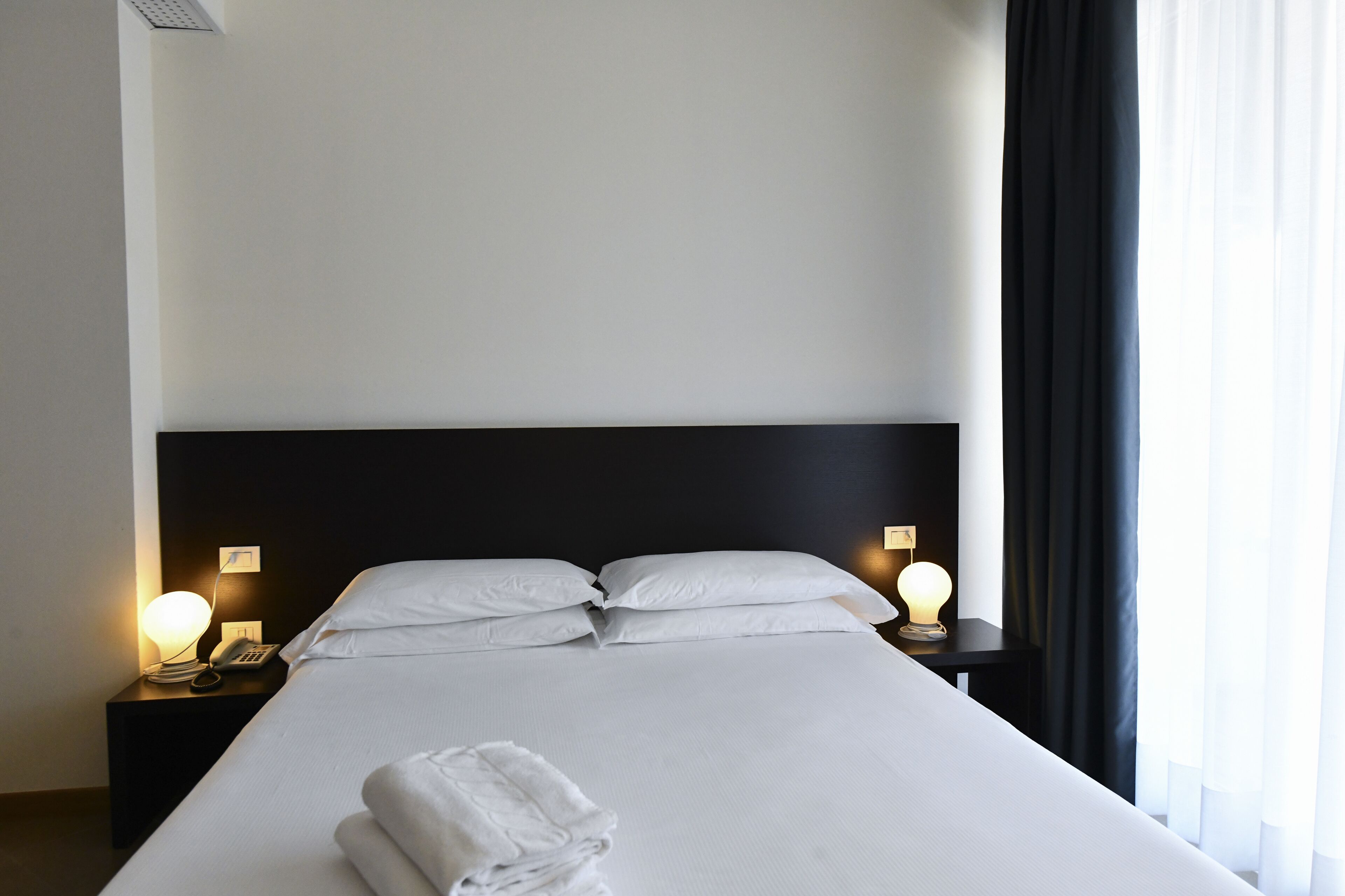 Una camera d'albergo con un letto rifatto, due lampade sui comodini, un telefono e un asciugamano piegato sul letto.