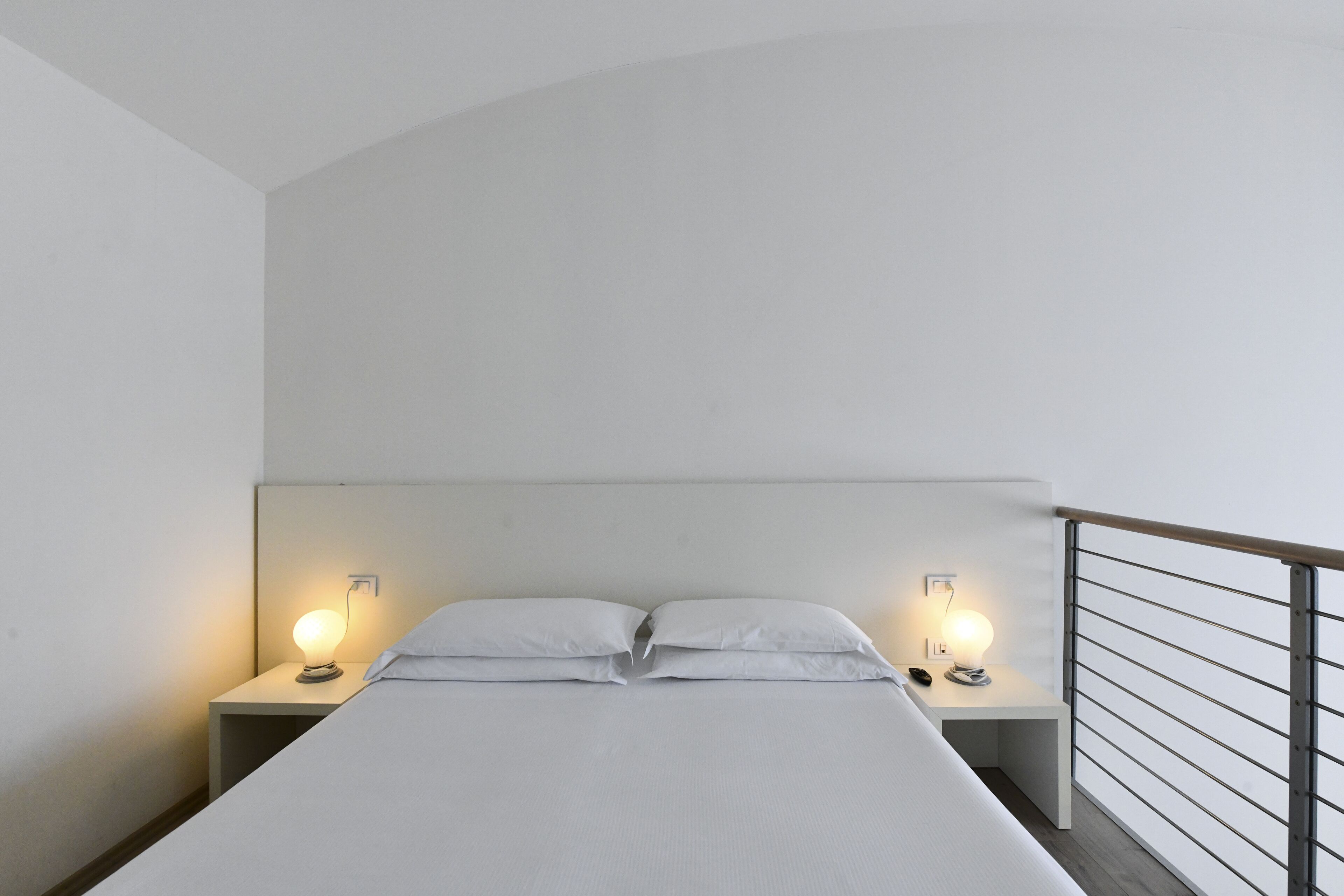 Un lit simple avec deux tables de chevet, chacune équipée d’une lampe, dans une pièce dont le plafond est en pente et qui possède une rambarde en bois.