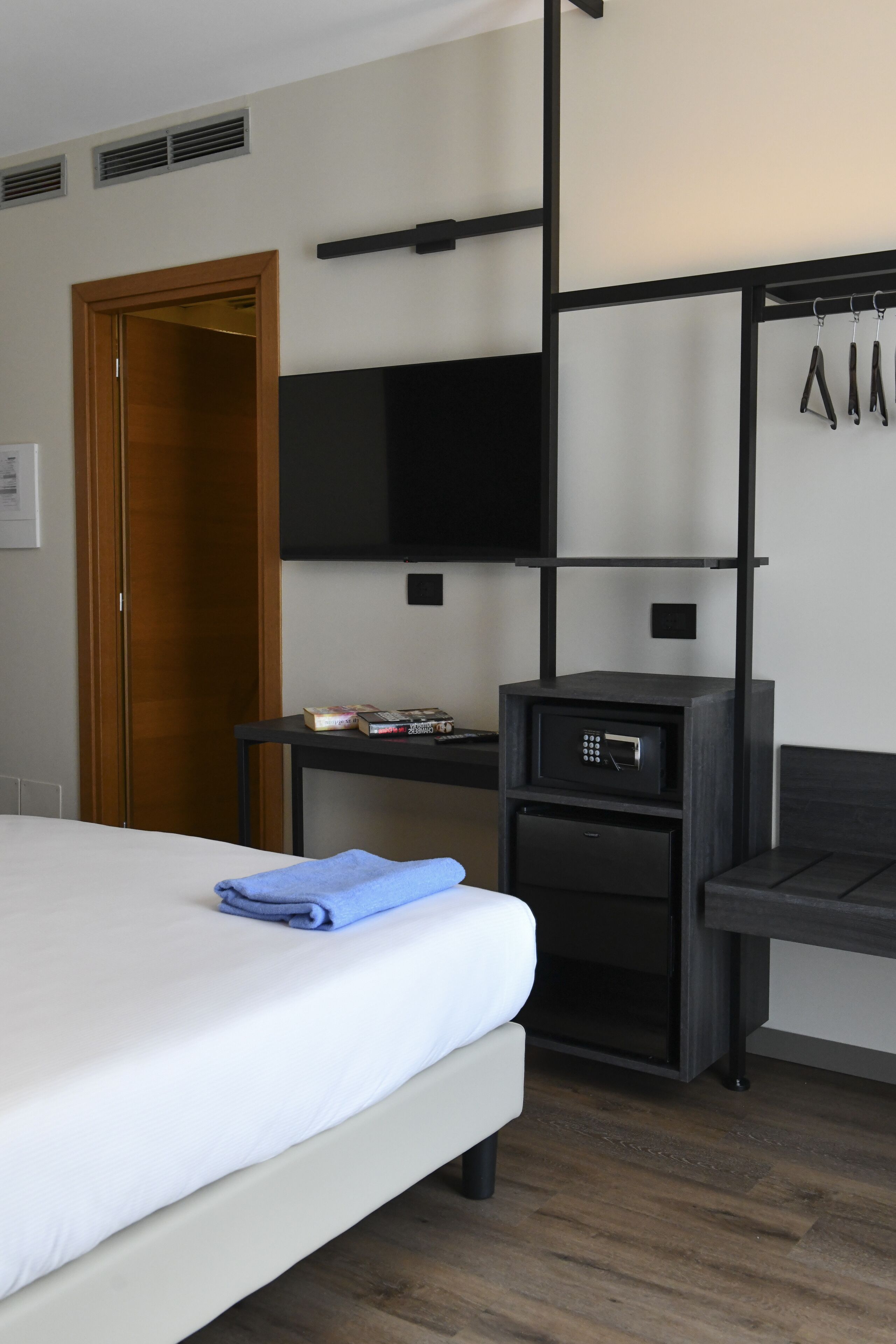 Ett hotellrum med en säng, en tv som är monterad på väggen, ett litet bord med ett värdeskåp och en klädställning.