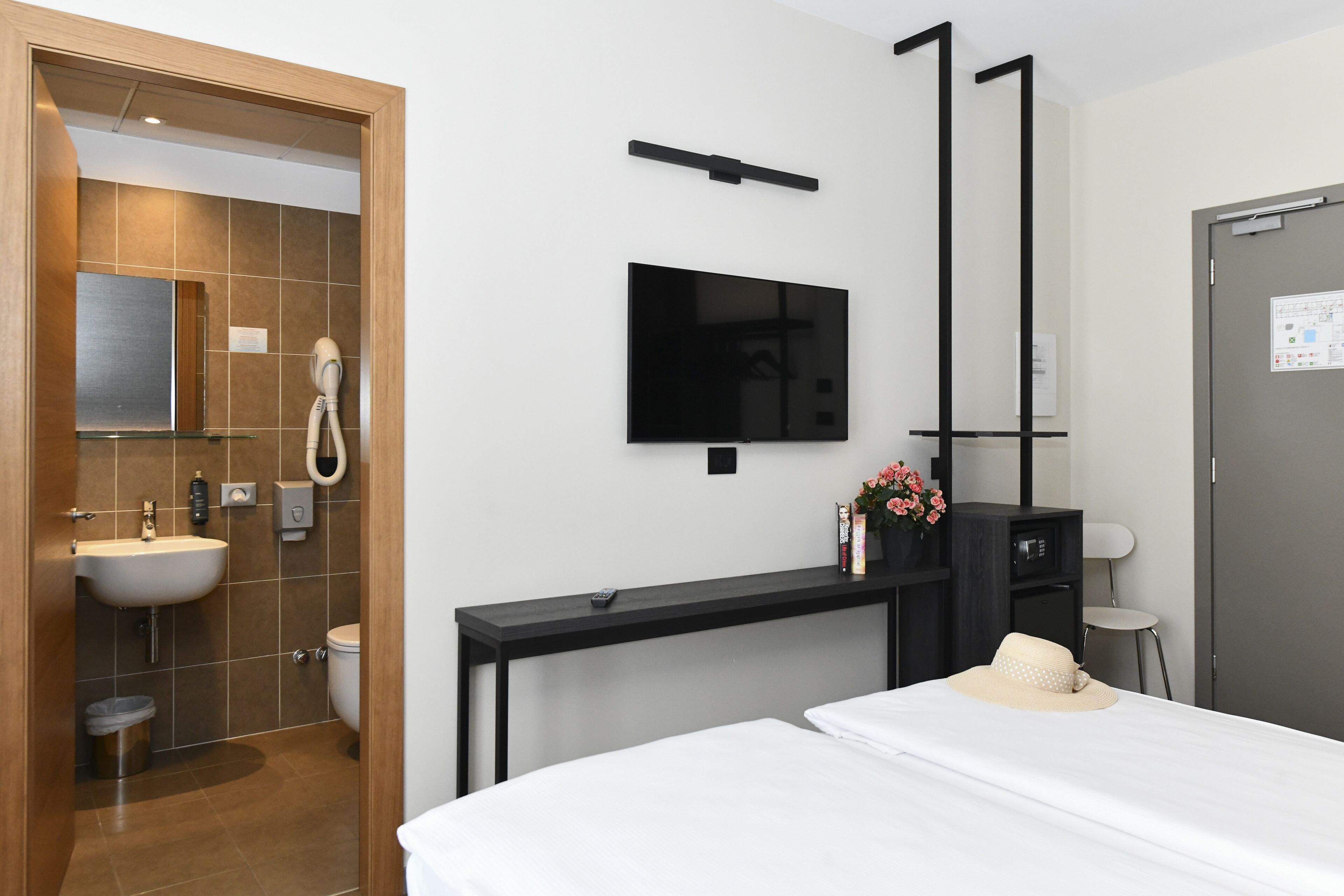 A modern hotel room with a bed, a desk, a TV mounted on the wall, a bathroom with a sink and toilet, and a door with a calendar.