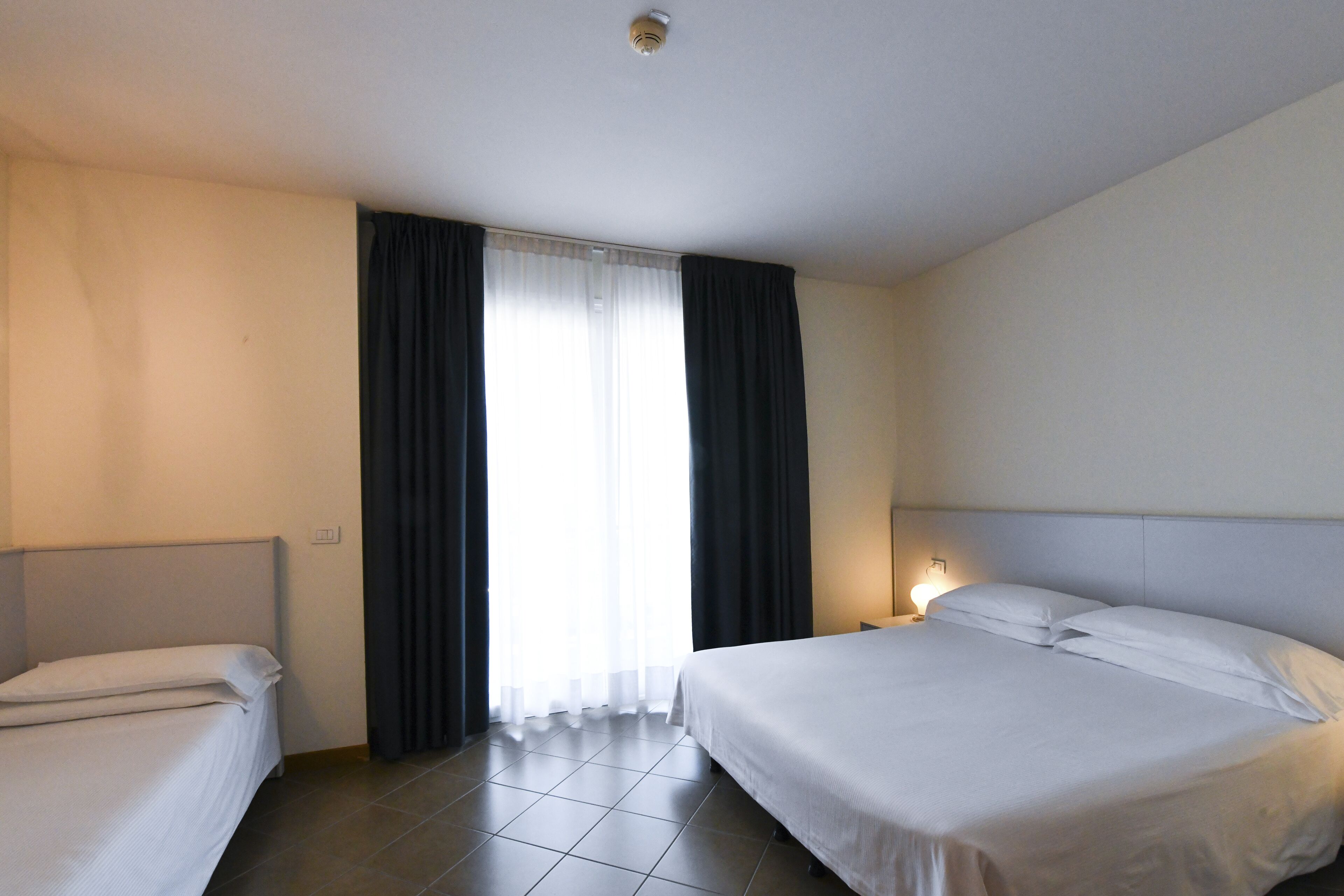 A hotel room with two beds, a window with curtains, a bedside lamp, and a tiled floor.