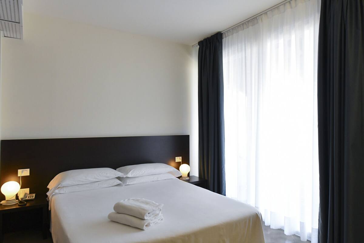 Una camera d'albergo con un letto rifatto, due lampade ai comodini e una finestra con tende.