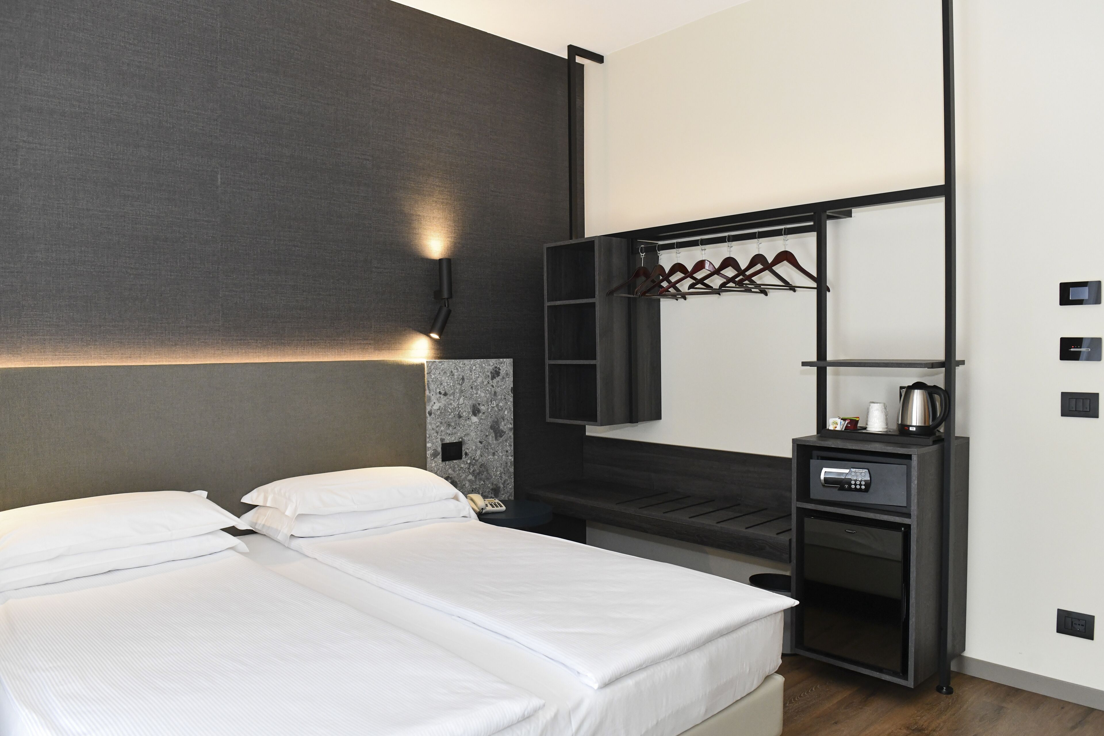 A modern hotel room with a bed, a bedside table with a coffee maker, a wardrobe with a mirror, and a wall-mounted light fixture.