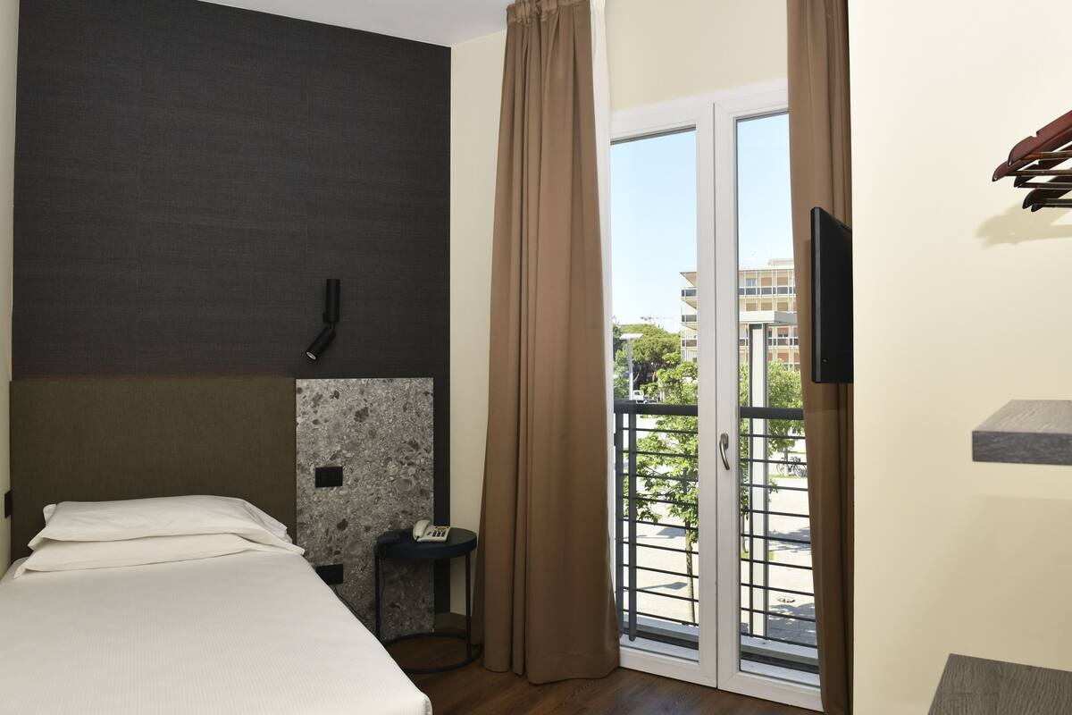 Camera d'albergo con un letto, un tavolino con un telefono, un telefono fissato al muro, una porta scorrevole in vetro che dà sul balcone e una TV montata a parete.