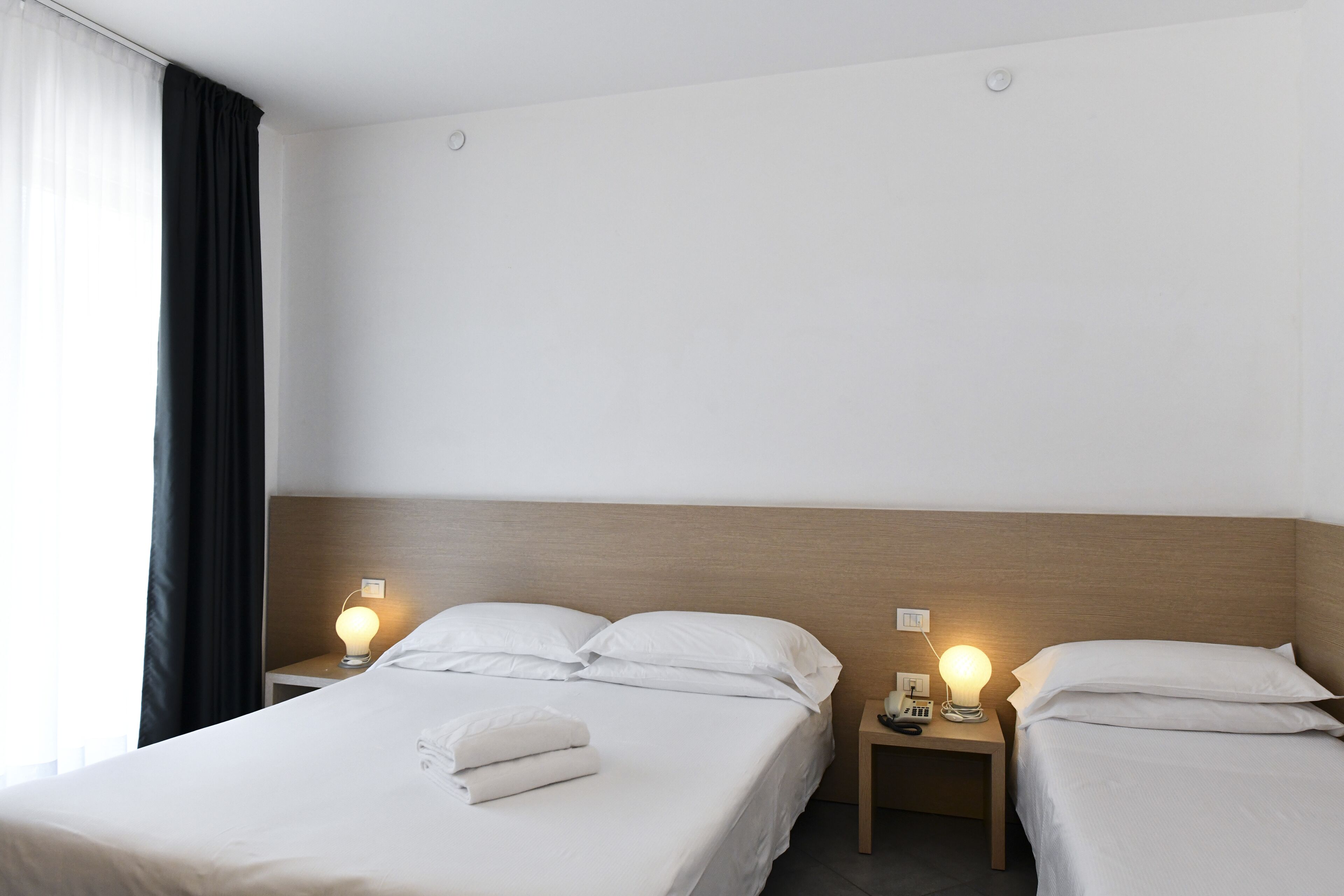 A hotel room with two beds, a wooden headboard, bedside tables with lamps, and a window with curtains.
